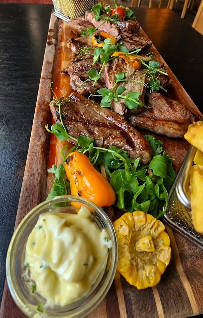 Tábua de madeira com fatias de carne grelhada, rúcula, pimentos pequenos e legumes grelhados, com um cesto de batatas fritas e um pote de vidro com creme claro