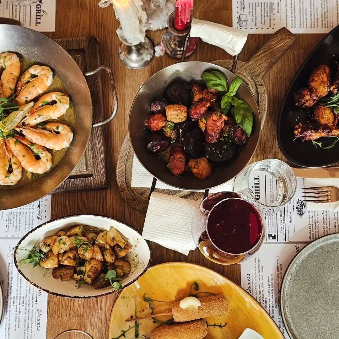 Mesa vista de cima com frigideira de camarões, tacho com chouriço e legumes assados, travessa de cogumelos, asas de frango, croquetes e um copo de vinho tinto