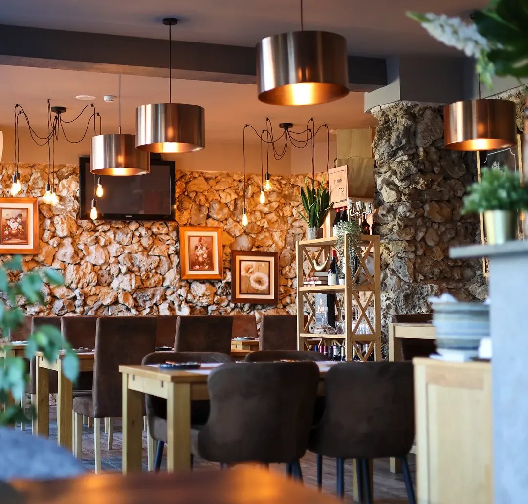 Sala com parede de pedra e quadros emoldurados, candeeiros de metal pendentes, mesas e cadeiras de madeira e plantas