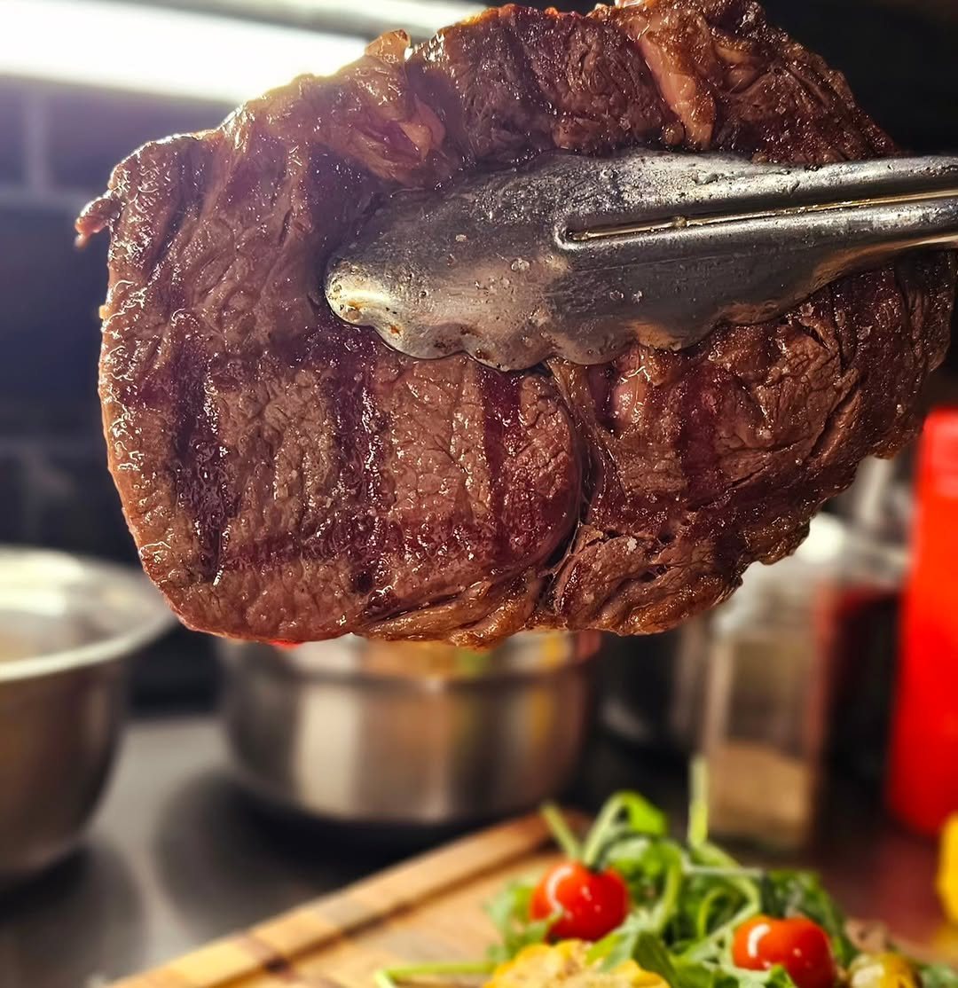 Pedaço de carne grelhada seguro com uma pinça, com salada de folhas e tomate-cereja em primeiro plano
